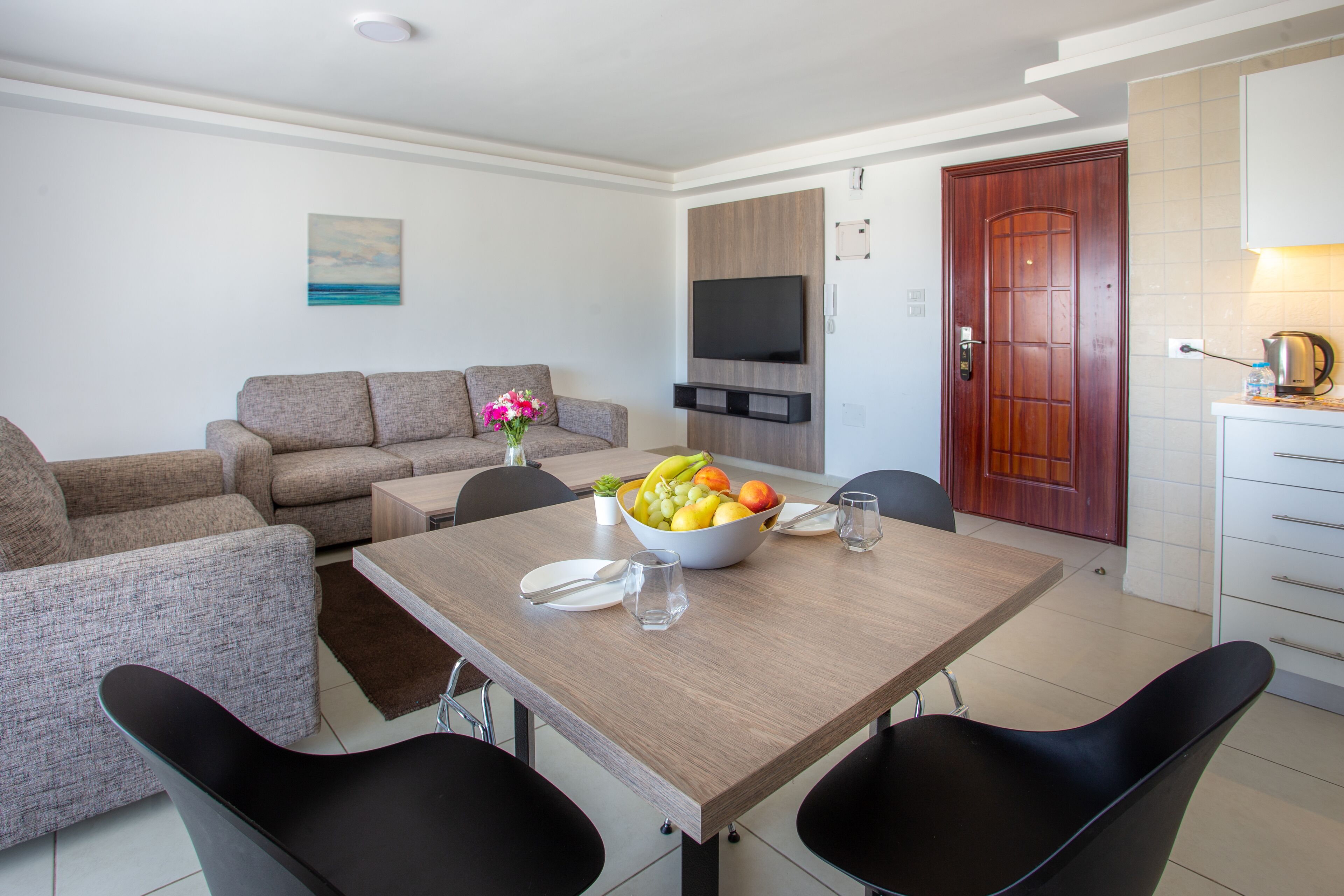 Una sala de estar moderna con un sofá, una mesa de comedor con sillas, un televisor montado en la pared y una puerta de madera.
