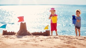 Deux enfants jouent avec du sable et des seaux sur une plage, près d’un château de sable et de drapeaux.