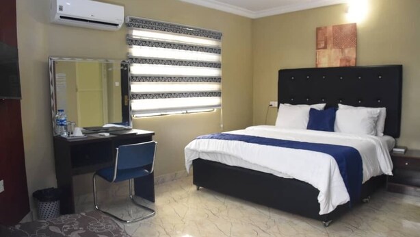 A hotel room with a bed, a desk with a chair, a mirror, and a window with blinds. - The Agore Hotels and Suites (Lekki)