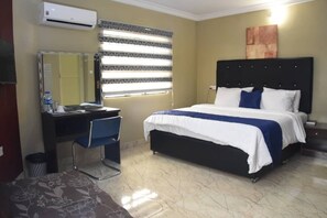 A hotel room with a bed, a desk with a chair, a mirror, and a window with blinds. - The Agore Hotels and Suites (Lekki)