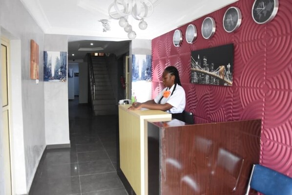 A reception area with a wooden desk, a receptionist, and a red patterned wall with clocks and cityscape artwork. - The Agore Hotels and Suites (Lekki)