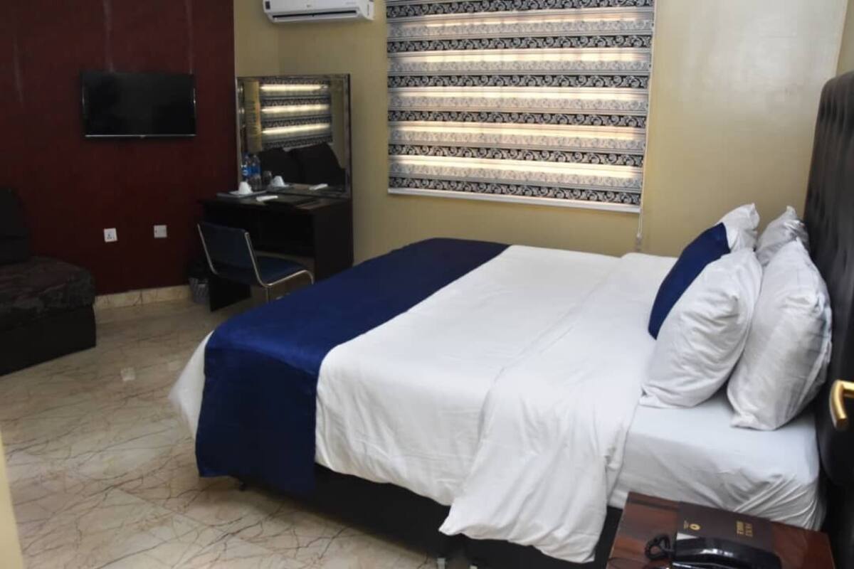 A hotel room with a bed, a desk with a computer, a chair, a TV mounted on the wall, and a window with patterned curtains.