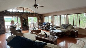 A spacious living room with a stone accent wall, a ceiling fan, and large windows offering a view of the outdoors. - Perfect relaxing lake retreat for hosting a gathering. Quiet with no neighbors. (Westminster)