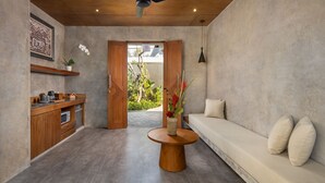 A modern interior with a wooden ceiling, a ceiling fan, a round wooden table, a sofa with cushions, and a door leading to an outdoor area with plants. - Agrapana Beach Villa (Sukawati)