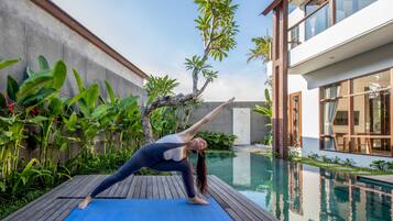 Una persona realizando una postura de yoga sobre una esterilla azul junto a una piscina, con una casa moderna y abundante vegetación al fondo.