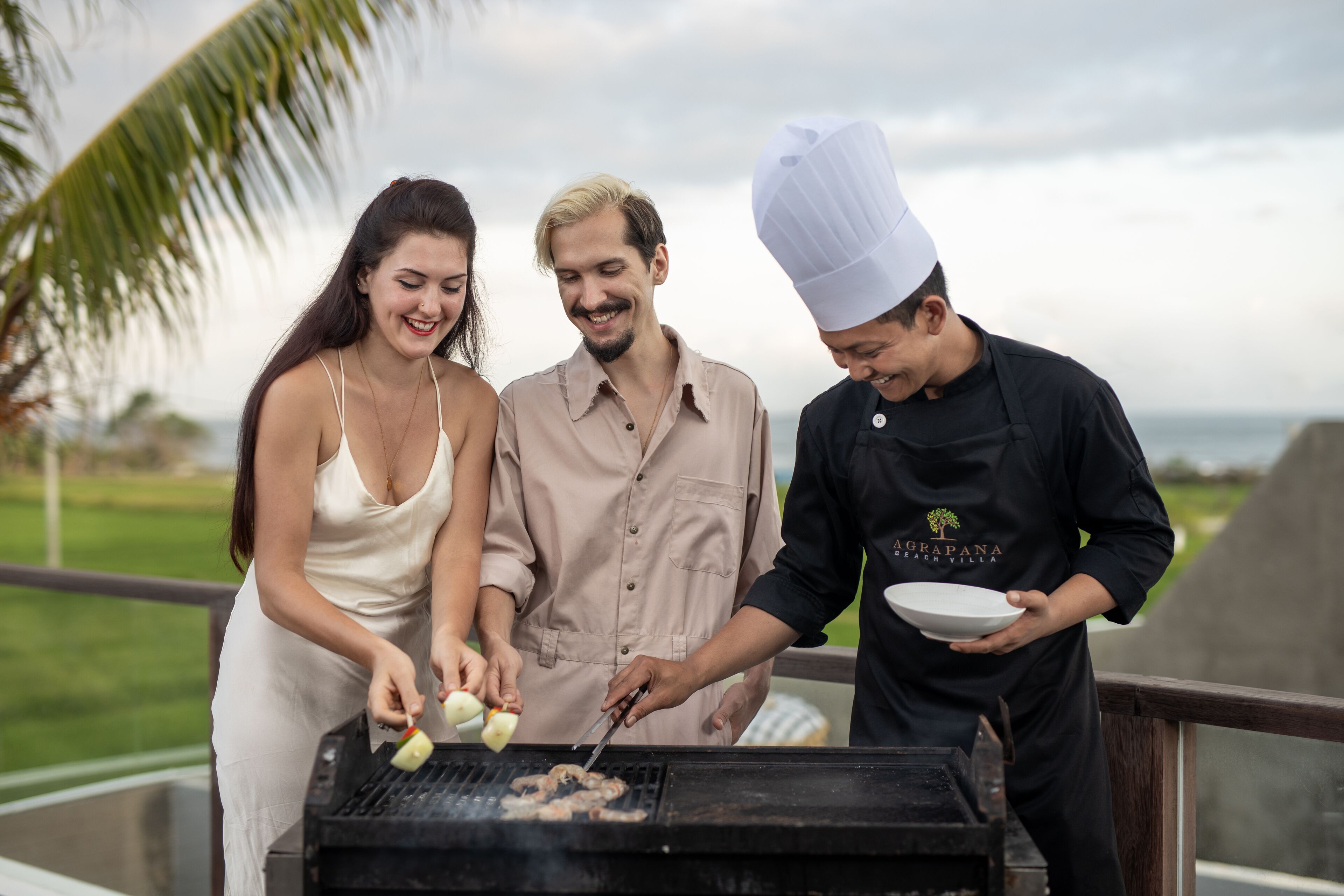 Three people grilling outdoors with a view of the ocean and greenery in the background.