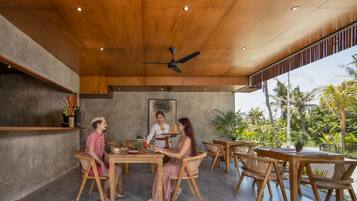 Techo de madera con iluminación empotrada, ventilador de techo y una mesa con sillas en una habitación con pared de hormigón y vistas a palmeras.