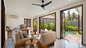 A modern living room with a sofa, coffee table, and a TV. There is a ceiling fan, a kitchen area with a stove, and a view of a garden through large sliding doors. - Agrapana Beach Villa (Sukawati)