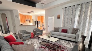 A modern living room with a grey sofa, armchairs, and a coffee table. There is a kitchen in the background with wooden cabinets and a dining area. - Remodeled luxury with Proximity (Lewisville)