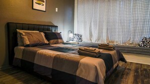 A neatly made bed with a striped bedspread and multiple pillows, a bedside lamp, and a window with a checkered curtain. - Remodeled luxury with Proximity (Lewisville)
