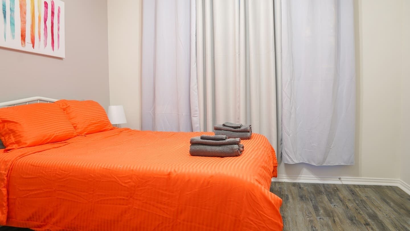 Une chambre d’hôtel avec un lit recouvert d’une literie orange, un mur blanc et une fenêtre avec des rideaux blancs.