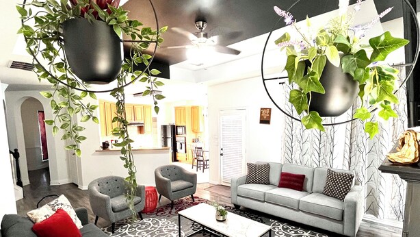 A modern living room with a grey sofa, red and white patterned rug, and a coffee table. There are two grey armchairs, a black floor lamp, and a hanging plant. The room has a large window with a view of the kitchen. - Remodeled luxury with Proximity (Lewisville)