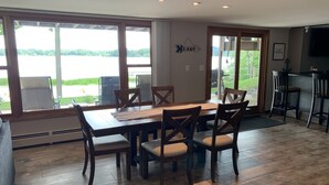 A dining area with a wooden table and chairs, large windows with a view of a lake, and a kitchen area with a bar counter. - Home w/ hot tub, game room, sauna on Lake Wausau near Granite Peak (Schofield)