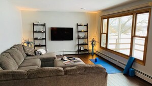 A living room with a sectional sofa, a flat-screen TV, two shelving units, and a large window with a view. - Home w/ hot tub, game room, sauna on Lake Wausau near Granite Peak (Schofield)