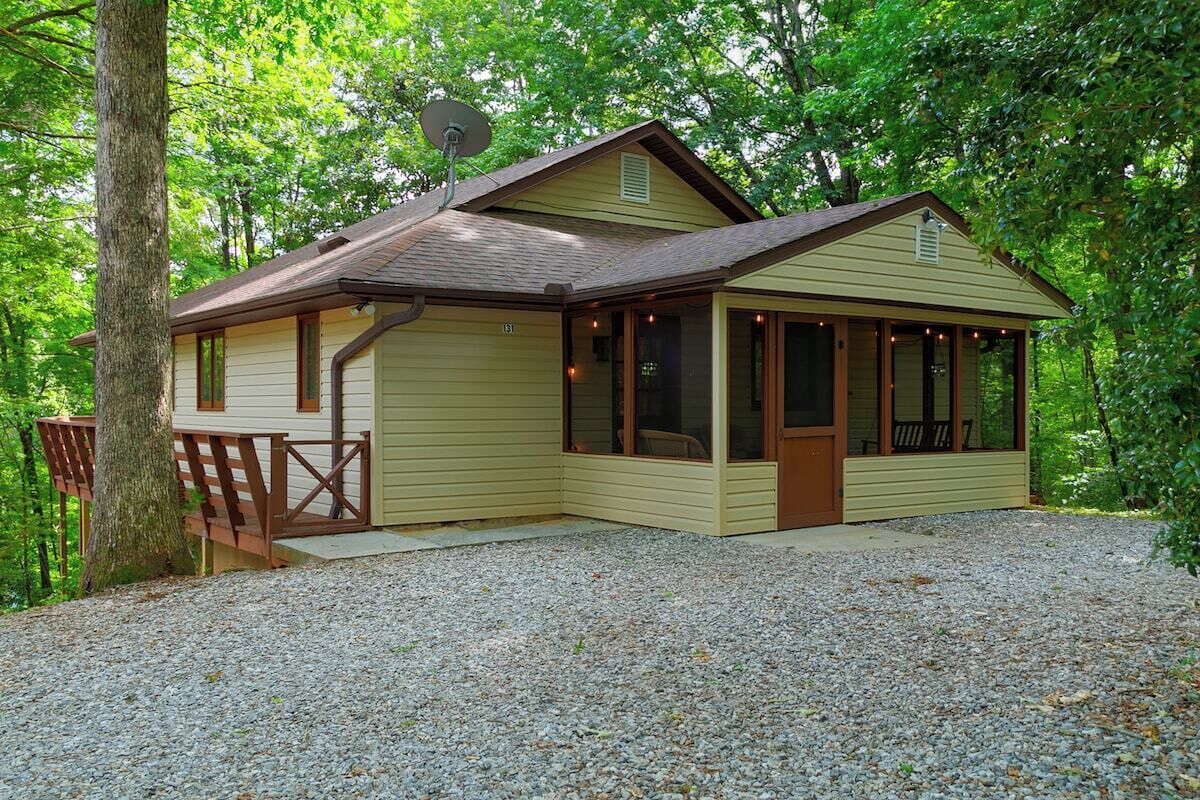 Une petite cabane à un seul étage, dotée d’un porche, d’une antenne parabolique et d’une allée en gravier, le tout entouré d’arbres.