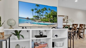 A living room with a flat-screen TV on a stand, a bookshelf with board games, a globe, and a potted plant.