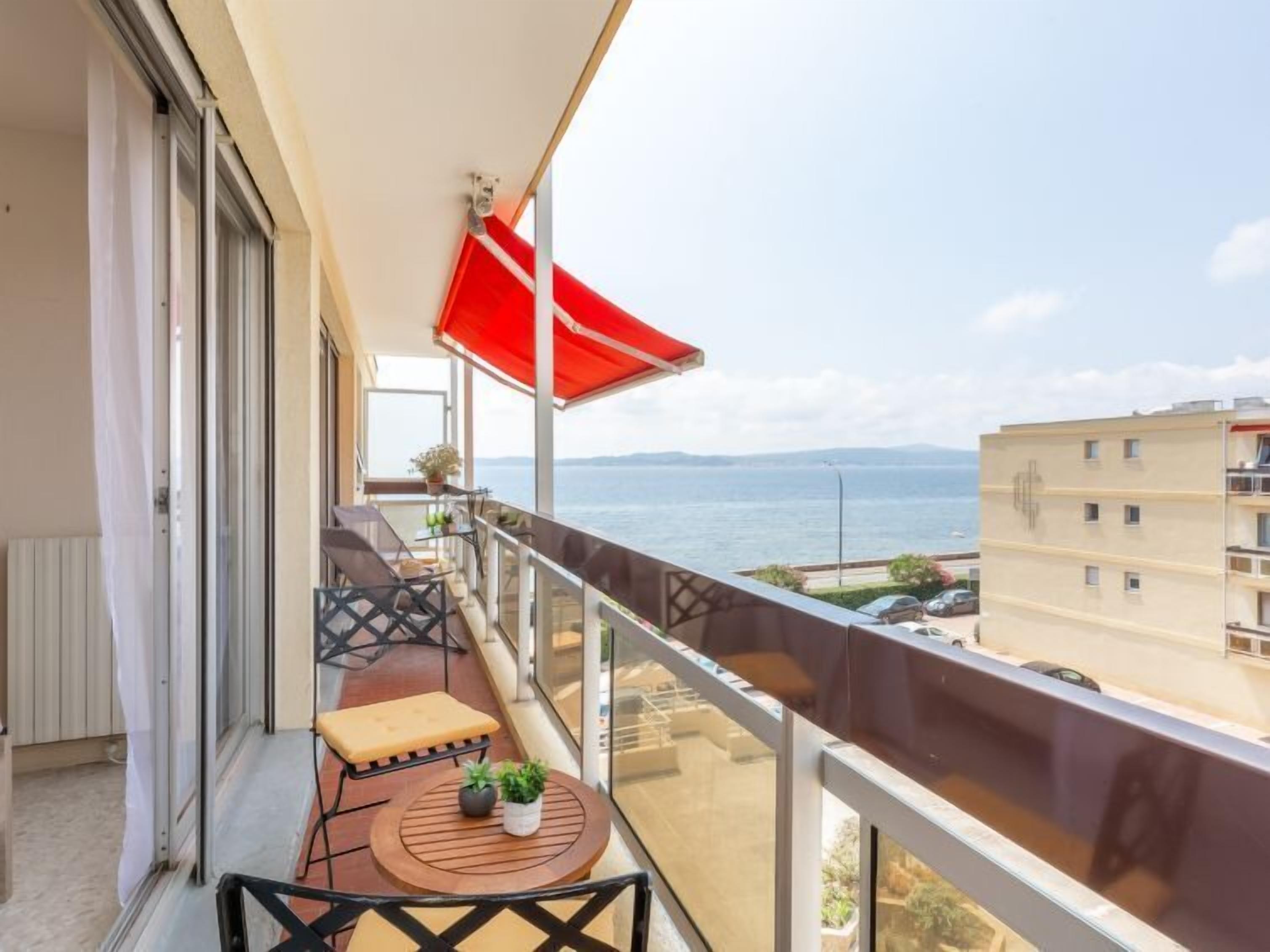 A balcony with a red umbrella, a table with two potted plants, a bench, and a view of the sea and buildings.