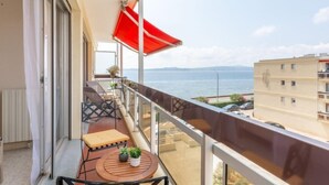Un balcone con un ombrellone rosso, un tavolo con due piante in vaso, una panchina e vista sul mare e sugli edifici.