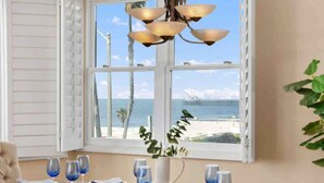 Une table à manger dressée pour six personnes, avec des verres à vin bleus, des assiettes et un pichet blanc, donnant sur une plage avec une jetée.