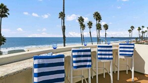 Une terrasse en bord de mer, avec des chaises et des tables à rayures bleues et blanches, d’où l’on peut admirer l’océan et les palmiers.