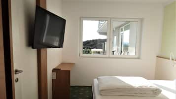 A hotel room with a bed, a television mounted on the wall, a window with a view of the outside, and a small wooden table.