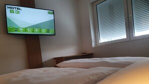 A hotel room with a bed, a TV mounted on the wall displaying the hotel's name and amenities, and a window with blinds. - Hotel B3 (Mauthausen)
