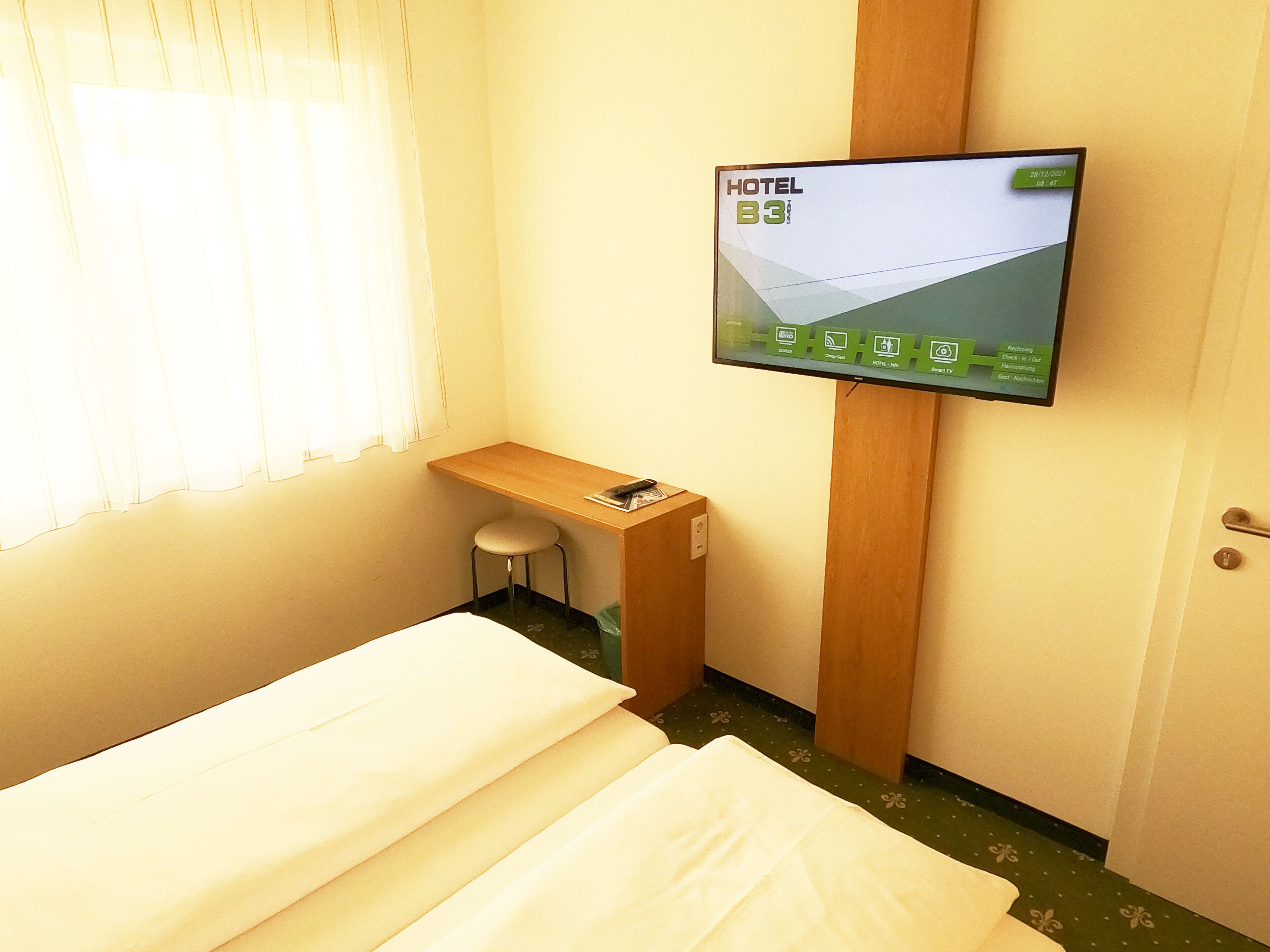 A hotel room with a bed, a small stool, a TV mounted on the wall, and a window with curtains.
