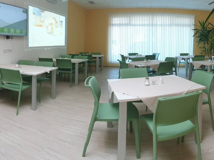 Zona de menjador amb cadires i taules verdes, una pantalla de projector a la paret i un gran finestral amb persianes.