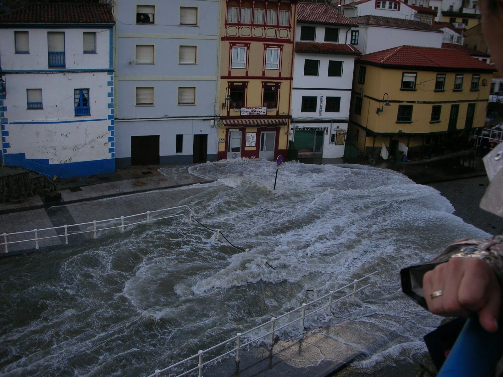 A view of a river with turbulent water, a railing, and buildings in the background.