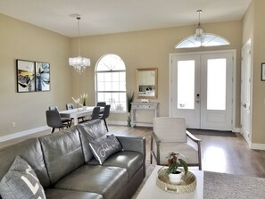 A spacious living room with a leather sofa, dining table, and chairs. There is a chandelier, a large window, and a door leading to another room. - Beautiful Vacation House Close to the Beach (Venice)