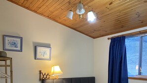 Une chambre avec un plafond en bois, un lit recouvert d’une couvre-lit à rayures grises et blanches, une table de chevet avec une lampe, et une fenêtre avec des rideaux bleus.