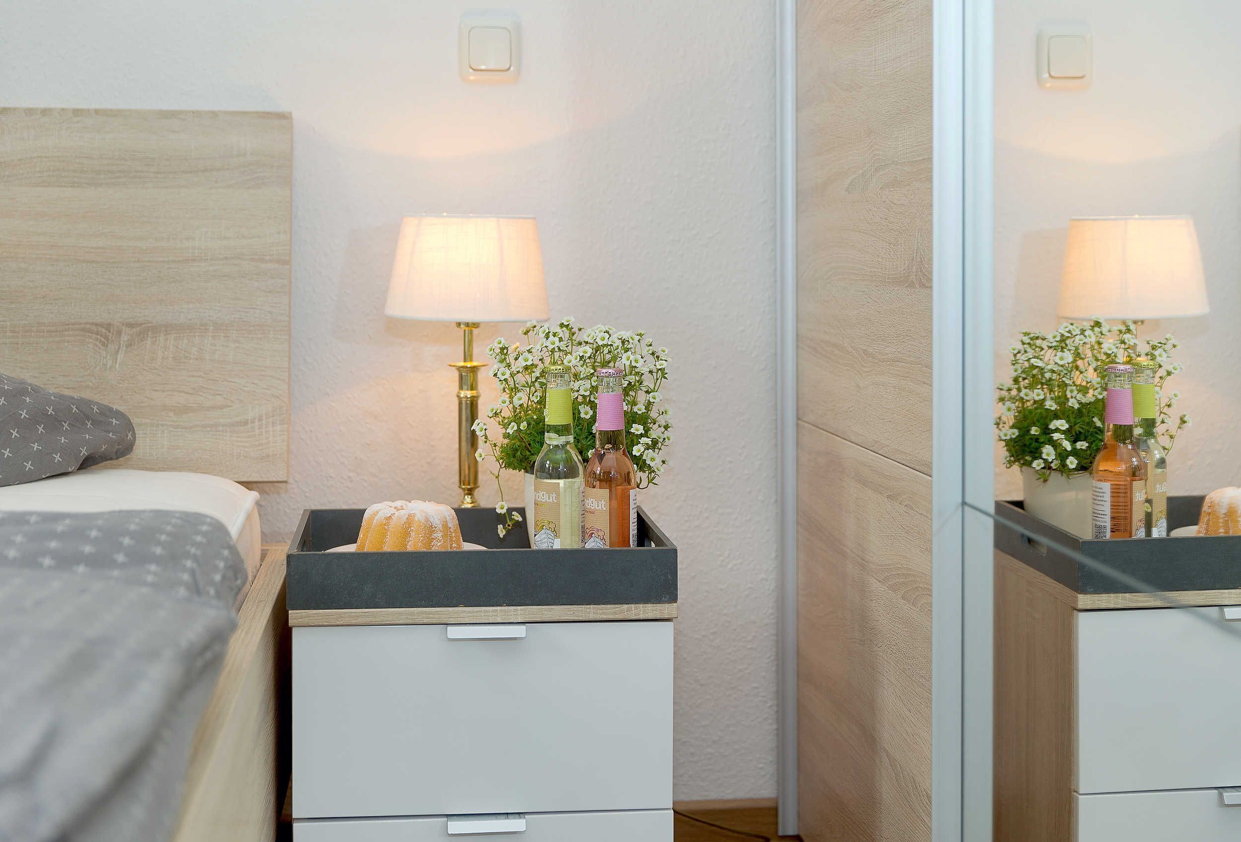 A neatly arranged bedroom with a bed, bedside tables, a mirror, and a nightstand with a lamp and flowers.