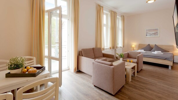 Una habitación de hotel moderna con sofá, sillón y cama. Hay una mesa de comedor con una bandeja de fruta y una planta pequeña.