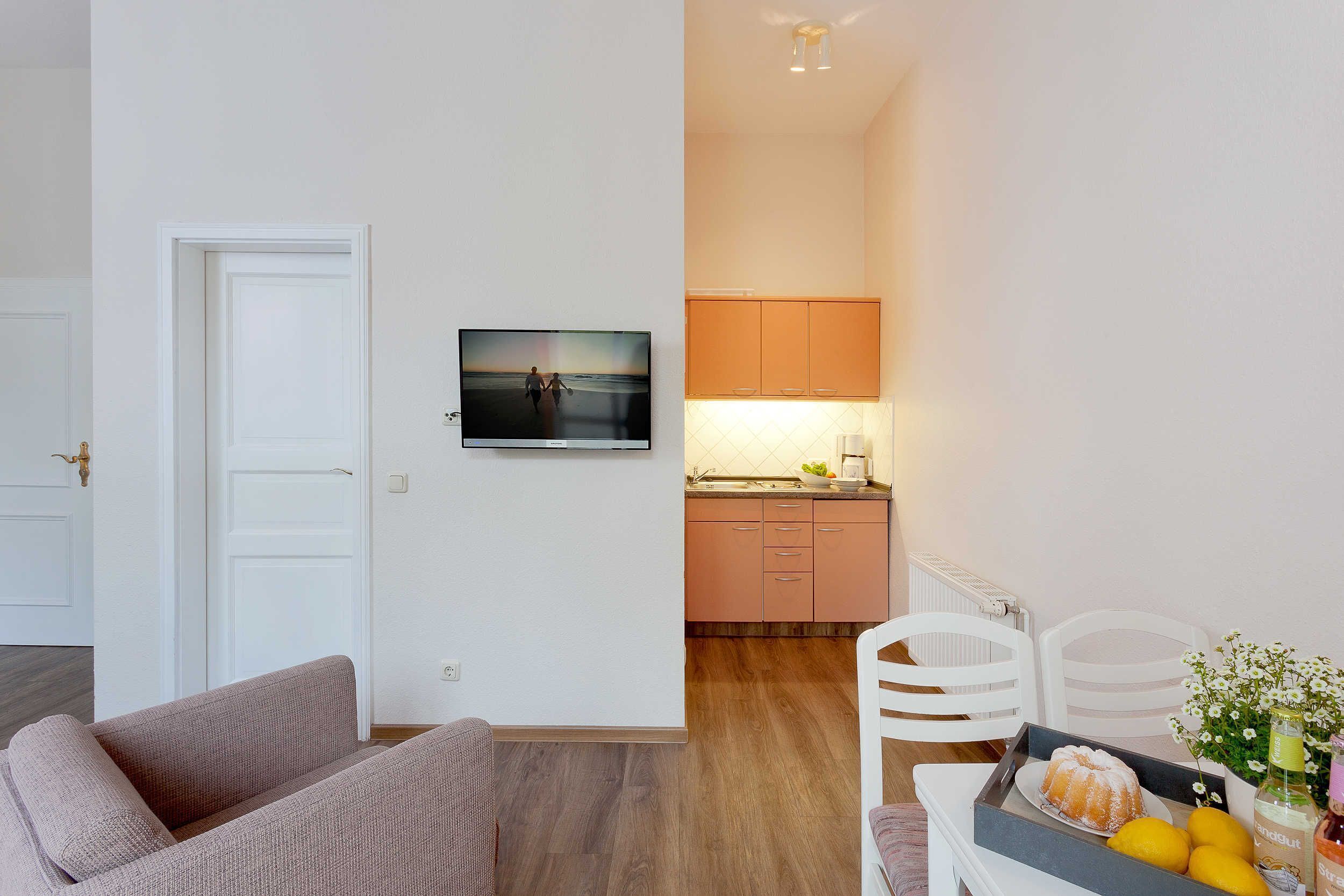 A modern living room with a sofa, a television mounted on the wall, and a kitchen area with cabinets and a countertop.