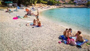 Une plage où des gens se prélassent au soleil et sont assis sur des serviettes, une mer bleue et limpide, et des bâtiments en arrière-plan.