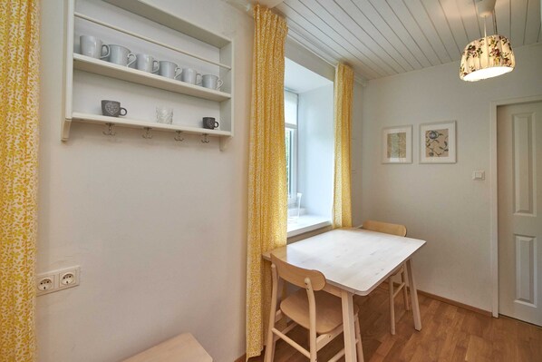 Una habitación con una mesa blanca, sillas de madera, un estante con tazas y una ventana con cortinas amarillas.
