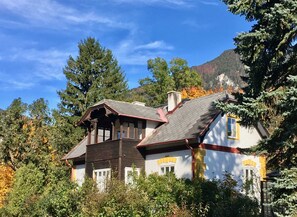 Una casa de dos pisos con un techo pronunciado, rodeada de árboles y una montaña al fondo.
