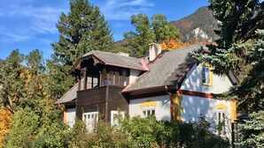 Une maison à deux étages, dotée d’un toit en pente, entourée d’arbres et avec une montagne en arrière-plan.