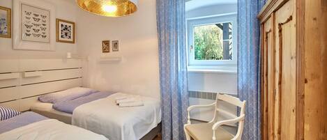A hotel room with two beds, a sitting chair, a wooden wardrobe, a window with curtains, and wall art.