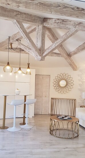 A room with exposed wooden beams, a white wall with a decorative mirror, a round metal table, and two white bar stools. - Studio Indépendant Villa (Mougins)