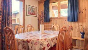 Een eetruimte met een ronde tafel, zes houten stoelen, een witte tafelkleed met rode patronen, een vaas met takken, en houten wandpanelen met een raam en gordijnen.