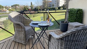 A balcony with wicker furniture, a table with plates and a wine bottle, and a view of a green lawn and buildings. - Elegant 3 rooms. Tennis pool and greenery. (Antibes)