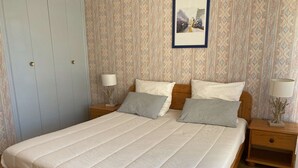 Una cama bien hecha con estructura de madera y ropa de cama blanca, dos mesitas de noche con lámparas, un papel tapiz estampado y una puerta con picaporte.