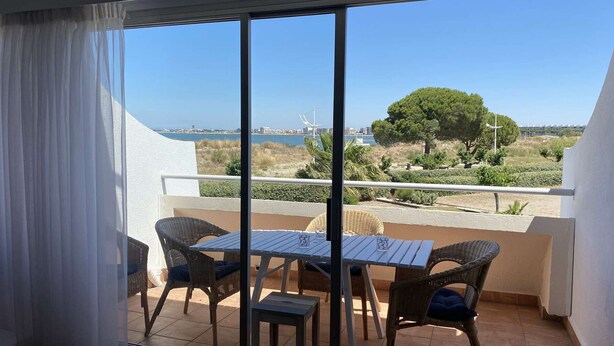 Un balcón con mesa y sillas, vista al mar y un árbol al fondo.