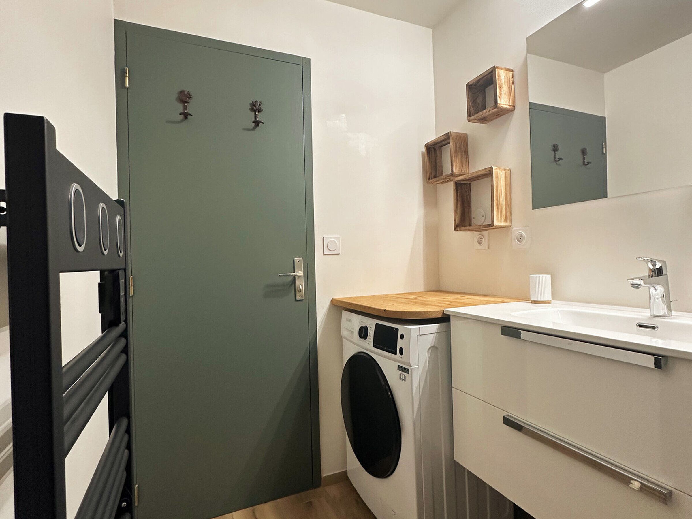 A compact, modern bathroom with a built-in washing machine, a sink with a mirror, and a wooden shelf.