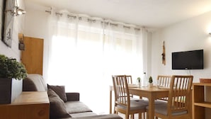 A cozy living room with a sofa, wooden dining table, and chairs. A television is mounted on the wall.