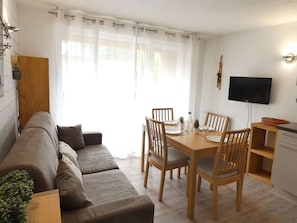 A cozy living room with a sofa, a dining table with chairs, a TV, and a small kitchen area.