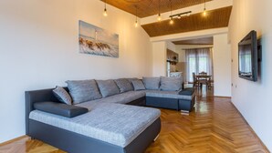 Een moderne woonkamer met een hoekbank, houten vloeren, een schilderij van een vuurtoren en een aan de muur bevestigde televisie.