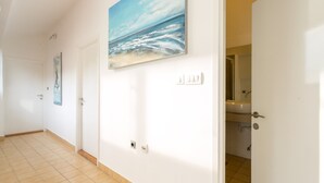 Een gang met een betegelde vloer, een schilderij van een strand en een badkamer zichtbaar door een open deur.