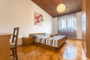 A bedroom with a wooden bed, a desk, and a chair. There is a window with curtains and a painting on the wall.